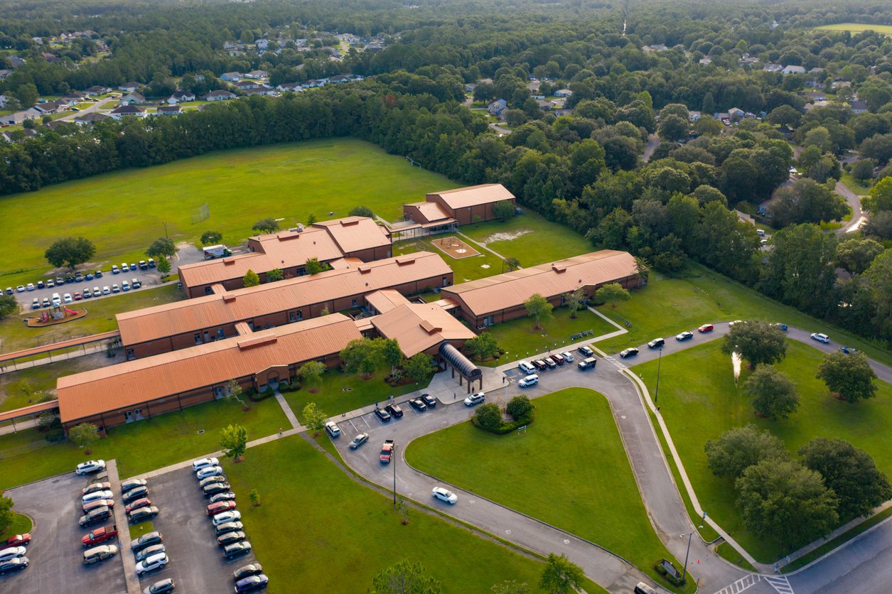 Aerial view of a large brick building with orange roofs surrounded by greenery and parking lots. Numerous cars are parked. Residential area in the distance.
